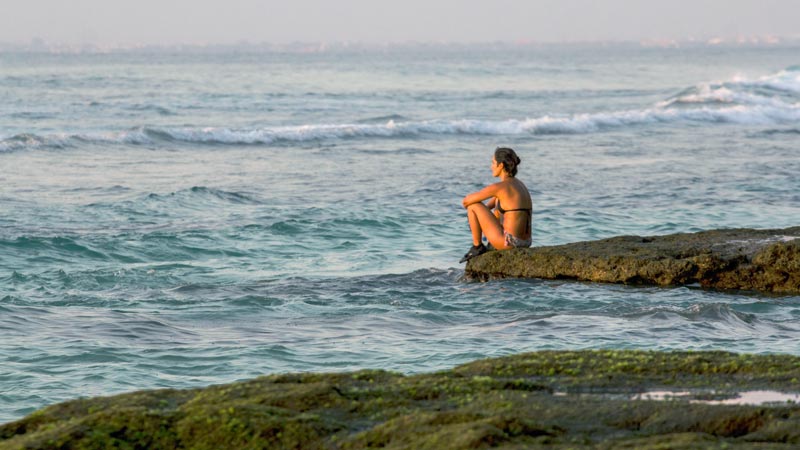 Pengunjung sedang duduk di batu karang di Pantai Suluban Uluwatu, menikmati pemandangan ombak dan laut biru.