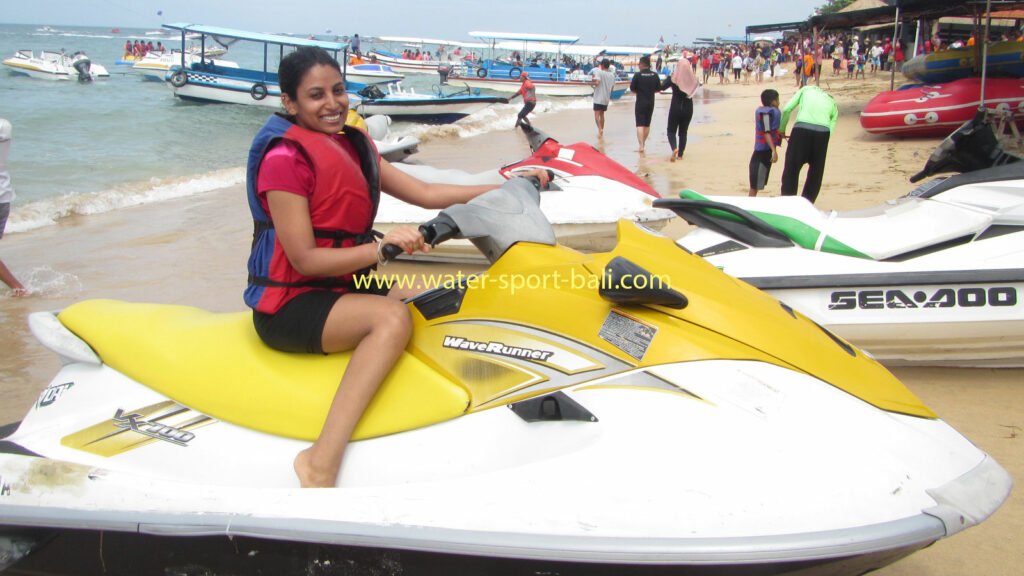 Seorang wanita tersenyum di atas jetski kuning, siap untuk petualangan air di Pantai Bali dengan mengenakan rompi keselamatan.