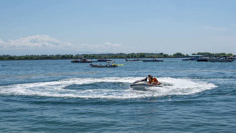 Wisatawan mengendarai jet ski dengan latar belakang perahu-perahu di Pantai Bali.