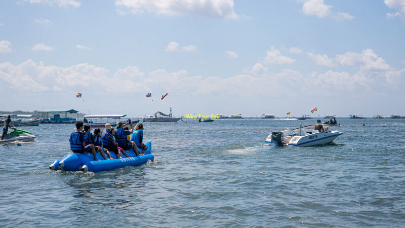 Wisatawan dengan jaket pelampung biru menaiki banana boat dengan latar belakang perahu dan parasut di Pantai