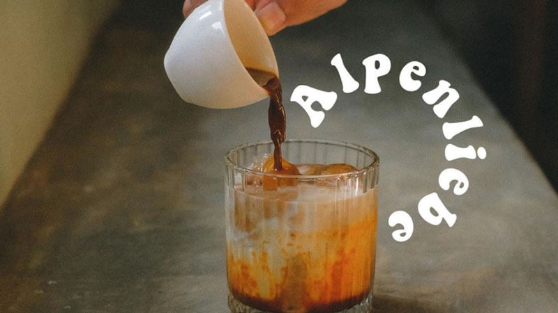 Tumpahan kopi susu segar ke dalam gelas es kopi dengan logo "Alpenliebe" di latar belakang.