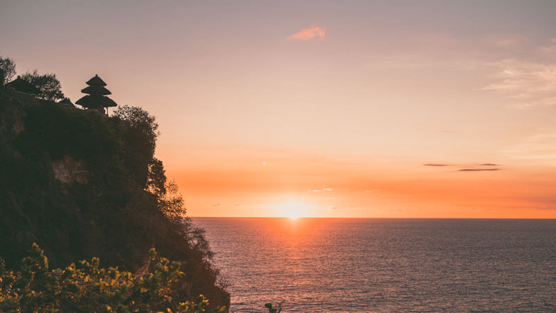 matahari terbenam dengan siluet pura di tepi tebing dan laut yang tenang di Bali