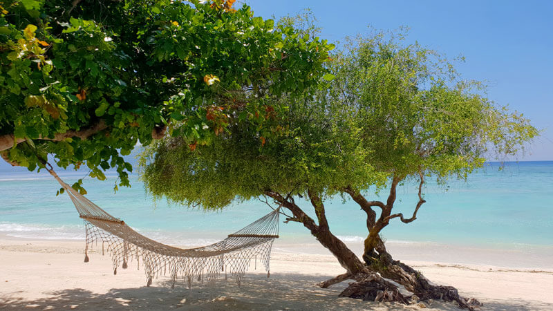 Hammock di bawah pohon rimbun dengan latar belakang pantai berpasir putih dan air laut biru di Kepulauan Gili T