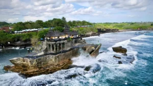 Pura Tanah Lot di atas batu karang di tengah laut, dikelilingi ombak dan pemandangan hijau di Bali.