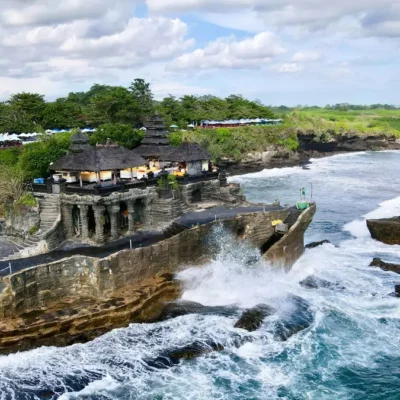 Pura Tanah Lot di atas batu karang di tengah laut, dikelilingi ombak dan pemandangan hijau di Bali.
