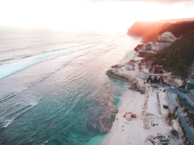 Foto aerial Pantai Melasti Ungasan saat matahari terbenam, menampilkan pasir putih, laut biru, dan jalan akses di antara tebing kapur.