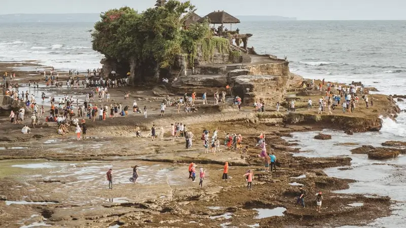 Pura Tanah Lot Bali dengan wisatawan yang berjalan di sekitar area pantai saat air surut, tempat wisata ikonik di Bali.