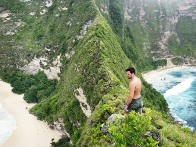Seorang traveler berdiri di tebing Kelingking Beach Nusa Penida, menikmati pemandangan laut dan pantai putih dari atas bukit ikonik.