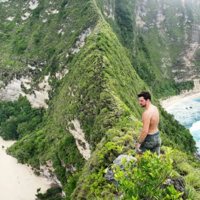Seorang traveler berdiri di tebing Kelingking Beach Nusa Penida, menikmati pemandangan laut dan pantai putih dari atas bukit ikonik.