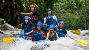 Grup peserta rafting melewati jeram dengan helm dan dayung di sungai.